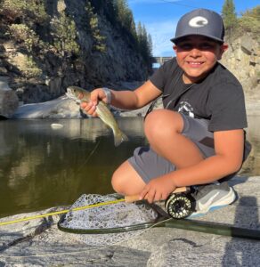A boy proudly holding a fish he caught by a river.